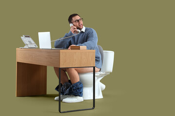 Young businessman on toilet bowl talking by telephone at table against green background - Powered by Adobe