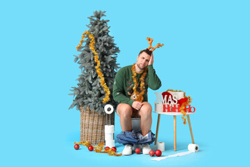 Young man sitting on toilet bowl with Christmas decor against blue background