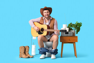 Young cowboy playing guitar on toilet bowl against blue background