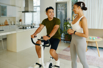 A man and woman discussing their workout routine while the woman uses a stationary bike in a bright living space