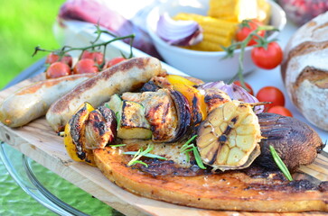 Close-up view of food being grilled on a backyard barbecue grill. The scene features sizzling sausages, juicy meat, and vibrant vegetables cooking over an open flame. 