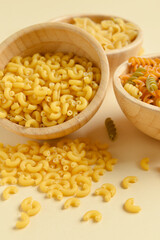 Wooden bowls with raw elbow and fusilli pasta on beige background, closeup