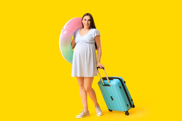 Young pregnant woman with inflatable ring and suitcase on yellow background. Travel concept