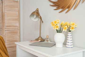 Vase with narcissus flowers, lamp and laptop on table in stylish room