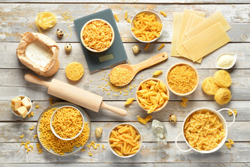 Different types of raw pasta and kitchen utensils on light wooden background