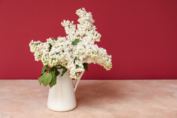Jug with beautiful blossoming lilac on table against red background