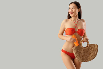 Young Asian woman in swimsuit with sunscreen cream and bag on light background