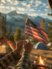 Person holding American flag in camping site with tent and campfire in scenic mountain landscape during sunset.