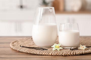 Glass and jug of fresh vanilla milk with sticks and flowers on wooden table in kitchen