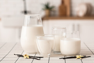 Glasses and jug of fresh vanilla milk with sticks and flowers on white tile table in kitchen