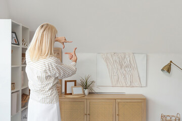 Mature woman looking at hanging 3D painting on light wall in living room, back view