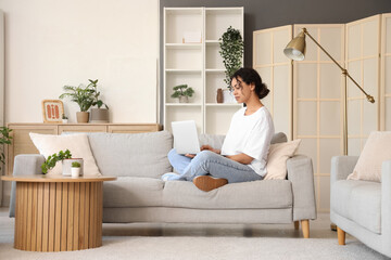 Young African-American woman using laptop on grey sofa at home
