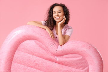 Beautiful young African-American woman with heart-shaped inflatable mattress on pink background