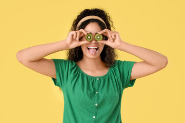 Beautiful young happy African-American woman with slices of kiwi on yellow background