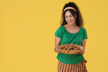 Beautiful young happy African-American woman with wicker plate full of kiwi on yellow background