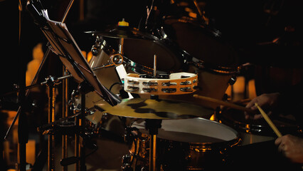 A drummer while playing drums. Focus on the drummer's hands holding the drumsticks in mid-movement. There are music sheets lying on a rack. Warm lighting emphasises ambience of a live performance.