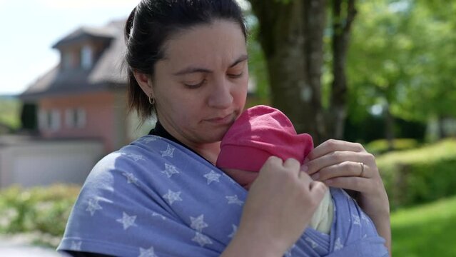Mother tenderly cradling newborn in sling while older son observes, capturing a moment of maternal affection, family bonding, and peaceful togetherness in an outdoor setting