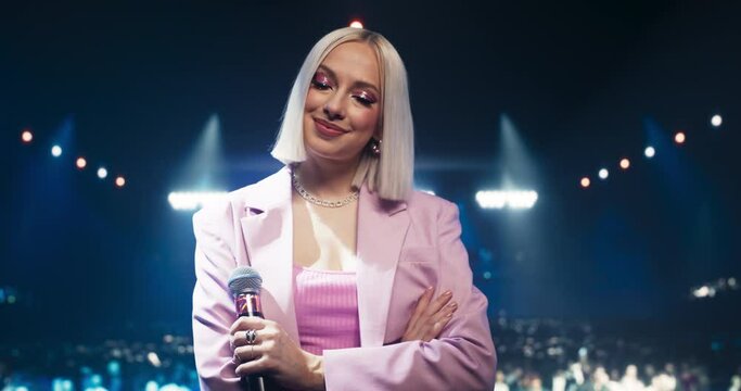 Portrait of a Young Blonde Female Idol Standing on Stage at a Concert, Posing in a Pink Outfit, Smiling and Looking at Camera. Successful Singer Standing with Her Arms Crossed and Holding a Microphone
