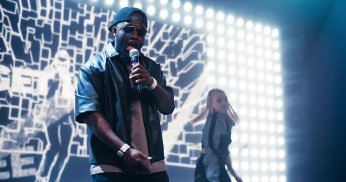 African American Hip Hop Artist Performing on Stage with a Dynamic Art Animation Background and a Female Dancer. MC Wearing Sunglasses and a Cap, Reading Energetic Rap Lyrics to the Audience