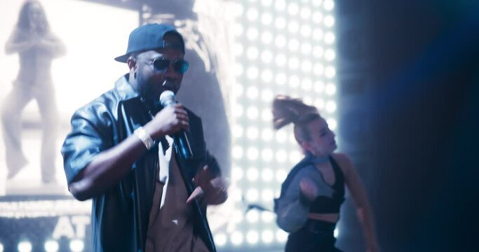 Gangster Rap Artist Performing on Stage with a Female Dancer that Makes Energetic Moves Under Spotlights. Creative Black Male Wearing Sunglasses and a Cap, Creating a Dynamic Concert Atmosphere