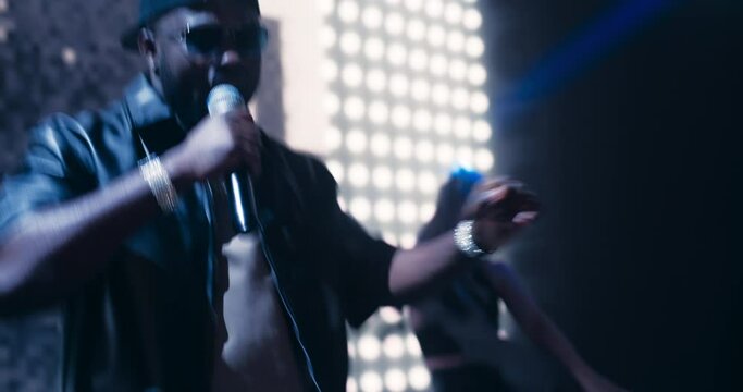 Hip Hop Concert with a Lively Performance: Young Talented Musician Performing a Show on Stage in Front of an Excited Audience of Fans and Music Lovers. Female Dancing Next to the African Artist