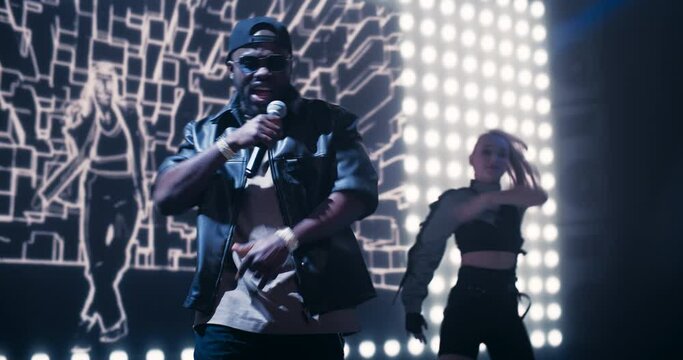 Young Hip Hop Singer in Black Leather Jacket Performing a Solo Concert with a Show Girl on a Festival Stage. His Charismatic Stage Presence and Lyrics Electrify the Diverse Music Fans in Crowded Arena