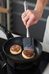 Hands of a female chef frying and baking homemade syrniki in a pan, sizzling in oil. Concept of home cooking, traditional recipes, and culinary preparation.
