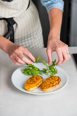Fish cutlets being placed on a plate and garnished with arugula salad and cherry tomatoes. Concept of home cooking, traditional healthy food recipes, and culinary presentation.