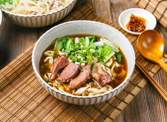 Classic Beef Tendon Noodles with chilli sauce, spring onion, wooden spoon, and chopsticks served in dish isolated on mat side view of taiwanese food