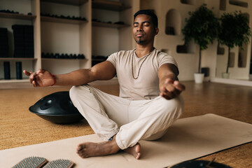 Portrait of mindful African American man meditating in lotus pose at home sitting on yoga mat with...