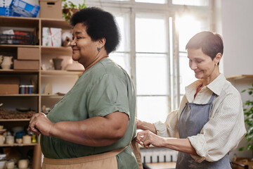 Side view portrait of two smiling adult women tying aprons preparing for art class in creative...