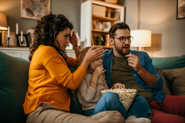 Family of three holding a popcorn pot and watching a scary movie together in the living room