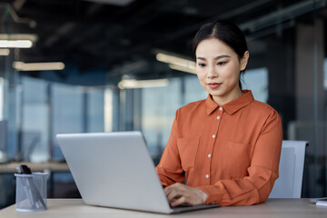 Serious thinking asian woman working inside office with laptop, business woman typing on keyboard, business woman financier preparing electronic report, report on company's financial results.