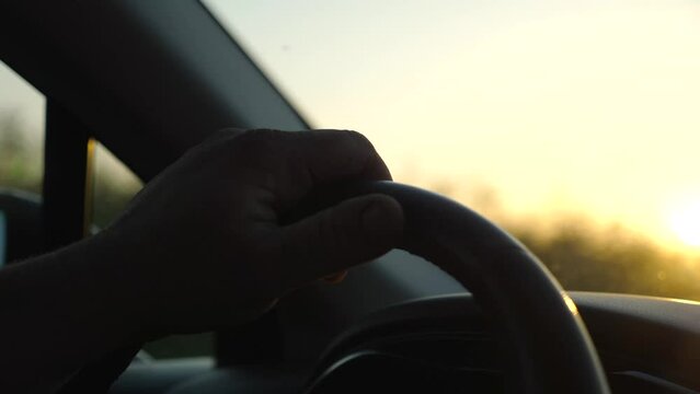 Male hand holds steering wheel while driving a motor car at sunset. Adult man operating the automobile on country road. Driver moving in his auto on highway. Concept of journey and trip. Close up