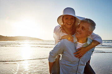 Mature, happy couple and piggyback with beach sunset for love, embrace or bonding together in nature. Man carrying woman with smile for hug, outdoor summer holiday or sunshine by water or ocean coast