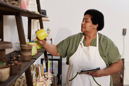 Side view portrait of senior African American woman inspecting glazed pottery pieces on shelf in art studio