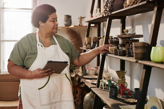 Side view portrait of Black senior woman inspecting pottery pieces on shelf in art studio lit by sunlight copy space
