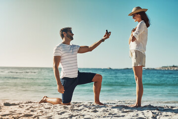 Love, engagement and couple on a beach, proposal and relationship for commitment, vacation and support. Surprise, man and woman on seaside holiday, sunshine and ask for hand, marriage with ring