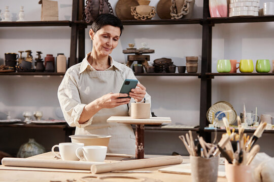 Waist up portrait of senior woman taking photos of handmade ceramics in art studio and posting on social media copy space