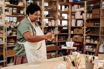 Side view portrait of African American senior woman taking photos of handmade ceramics via smartphone in pottery studio copy space