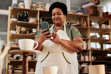 Low angle portrait of African American senior woman using smartphone in pottery studio and posting pictures on social media copy space