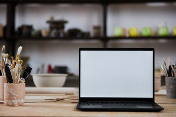 Background image of open laptop with blank white screen on wooden table in pottery studio, copy...