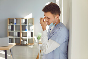 Portrait of stressed male doctor wearing blue uniform and face mask standing in hospital suffering from headache. Young exhausted man physician or nurse tired from hard work in medical clinic.
