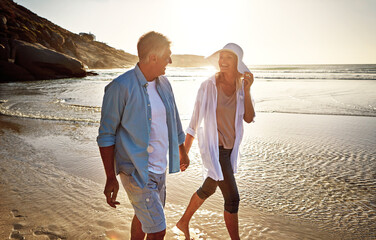 Mature, happy couple and walking with sunset on beach for love, embrace or bonding together in nature. Man, woman or lovers holding hands with smile for summer holiday by water, sea or ocean coast