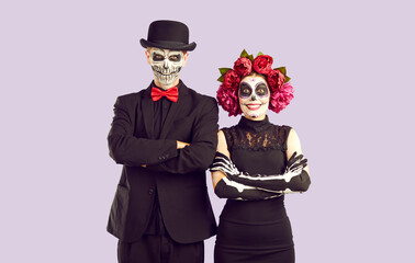 Adorable zombie girl in peony wreath and her boyfriend with skull on his face celebrate Halloween. Portrait of cheerful smiling couple standing with folded hands on lilac background. Halloween concept