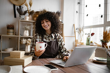 Young Caucasian woman potter happy with online sale of mug on Internet against background of...