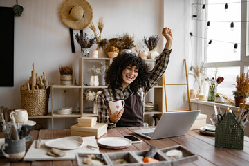 Young Caucasian woman potter happy with online sale of mug on Internet against background of...