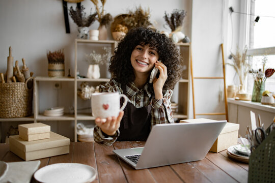 Successful business lady owner of shop made of clay talking to customer. Caucasian female potter entrepreneur talking to buyer of handmade mug sitting at table and taking order using laptop. - Powered by Adobe