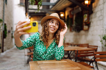Young woman holding a smartphone digital camera with her hands and taking a selfie while sitting at the summer cafe. Fashion, beauty, blogging, tourism.