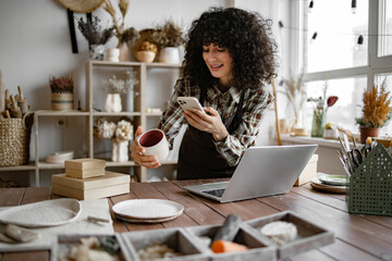 Successful girl store employee with ceramic tableware shoots photos on smartphone. Charismatic...